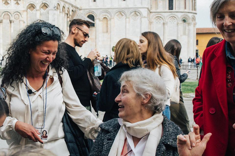 Group of people smiling and talking around a café table with coffee cups in front of Pisa Cathedral, Italy.