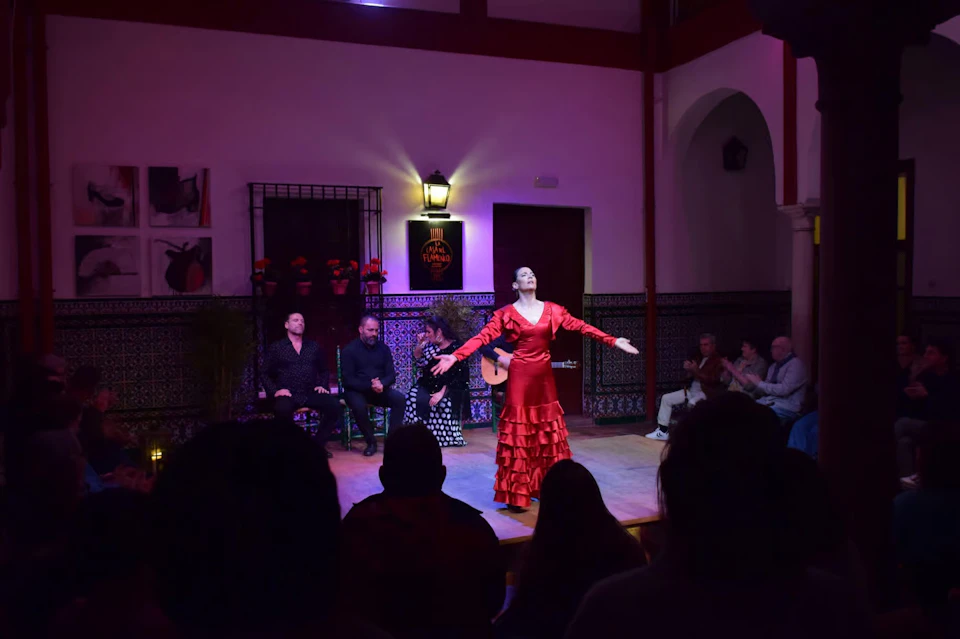 A flamenco dancer in a red dress performs on a stage with musicians seated behind her, in front of an audience.