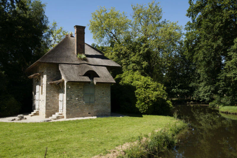 A small stone house with a thatched roof next to a river, surrounded by trees and greenery on a sunny day.