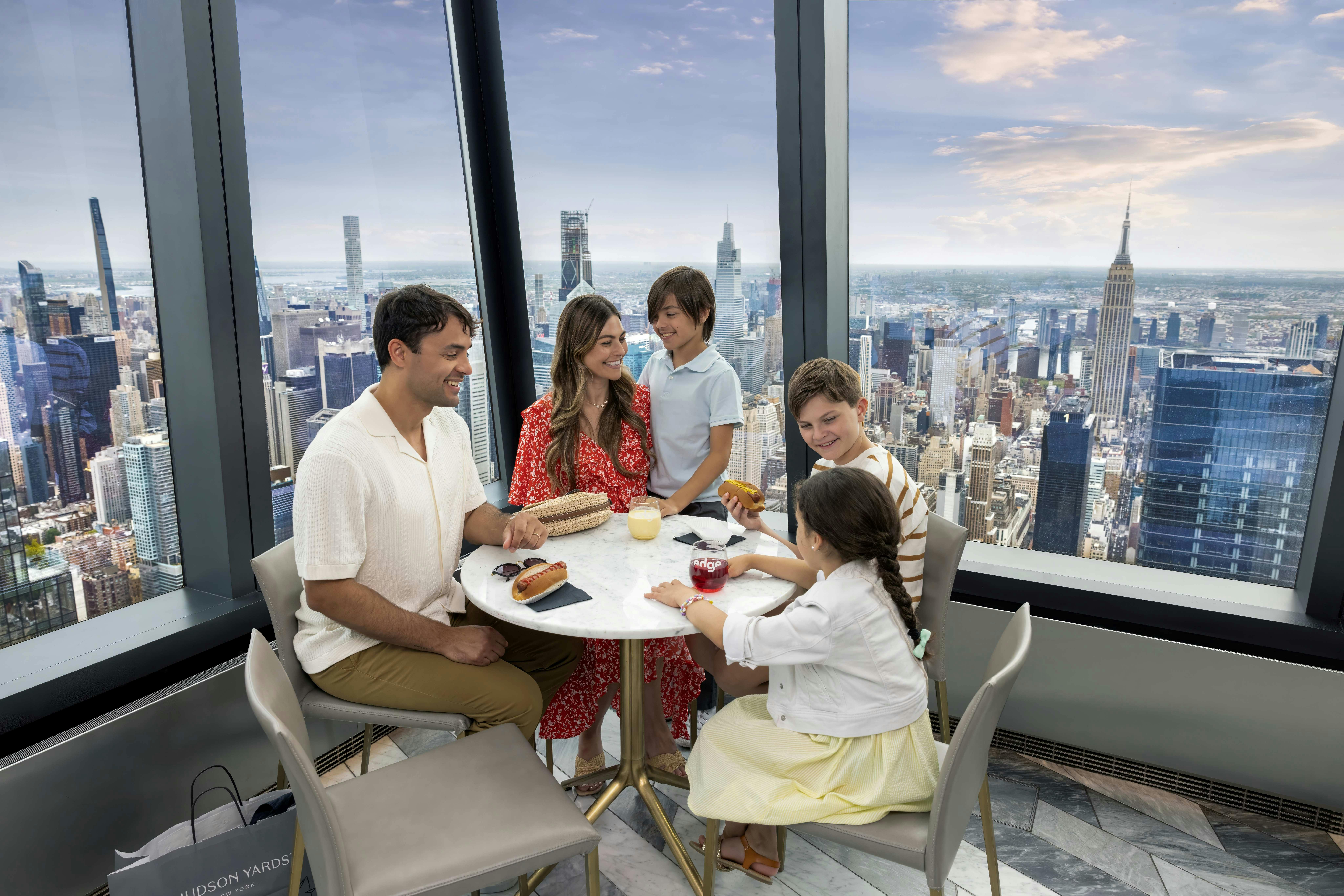 5인 가족이 도시 스카이라인이 내려다보이는 커다란 창문 옆 테이블에 앉아 음식과 음료를 함께 즐기고 있습니다.