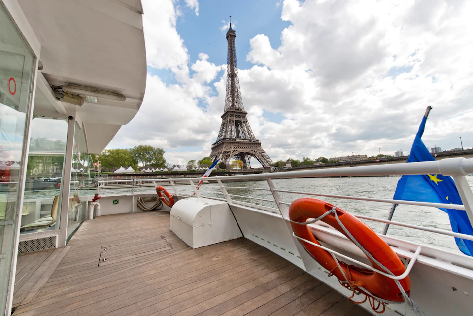 View of the Eiffel Tower from the deck of a boat on the Seine River, featuring life buoys and wooden flooring under a partly cloudy sky.