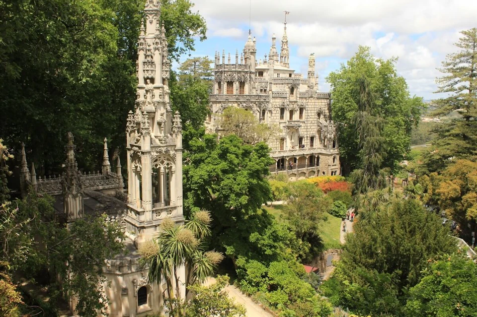 Historic, ornate building with spires surrounded by lush greenery and gardens on a sunny day.