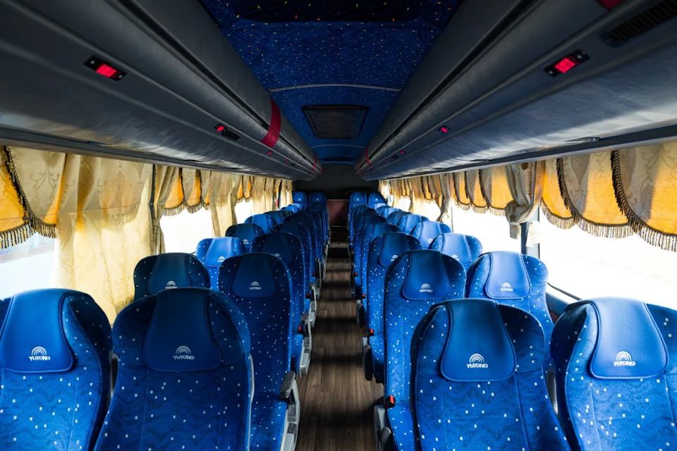 Interior of a bus with rows of blue seats featuring a dotted pattern, yellow curtains along the windows, and aisle in the center.