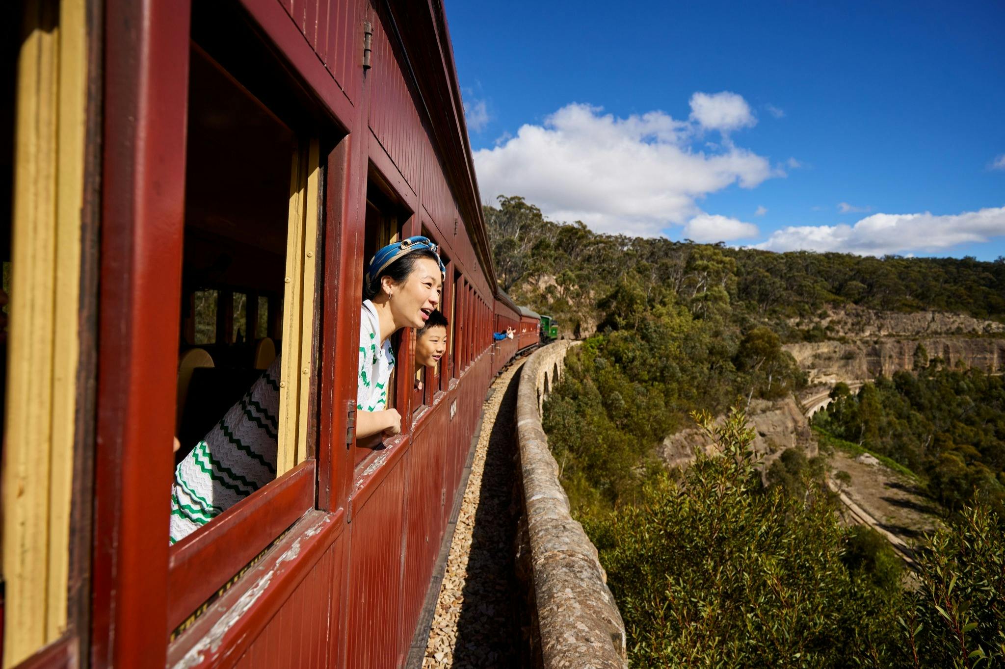 Two people lean out of windows of a red train traveling along a scenic route with trees and blue sky in the background.