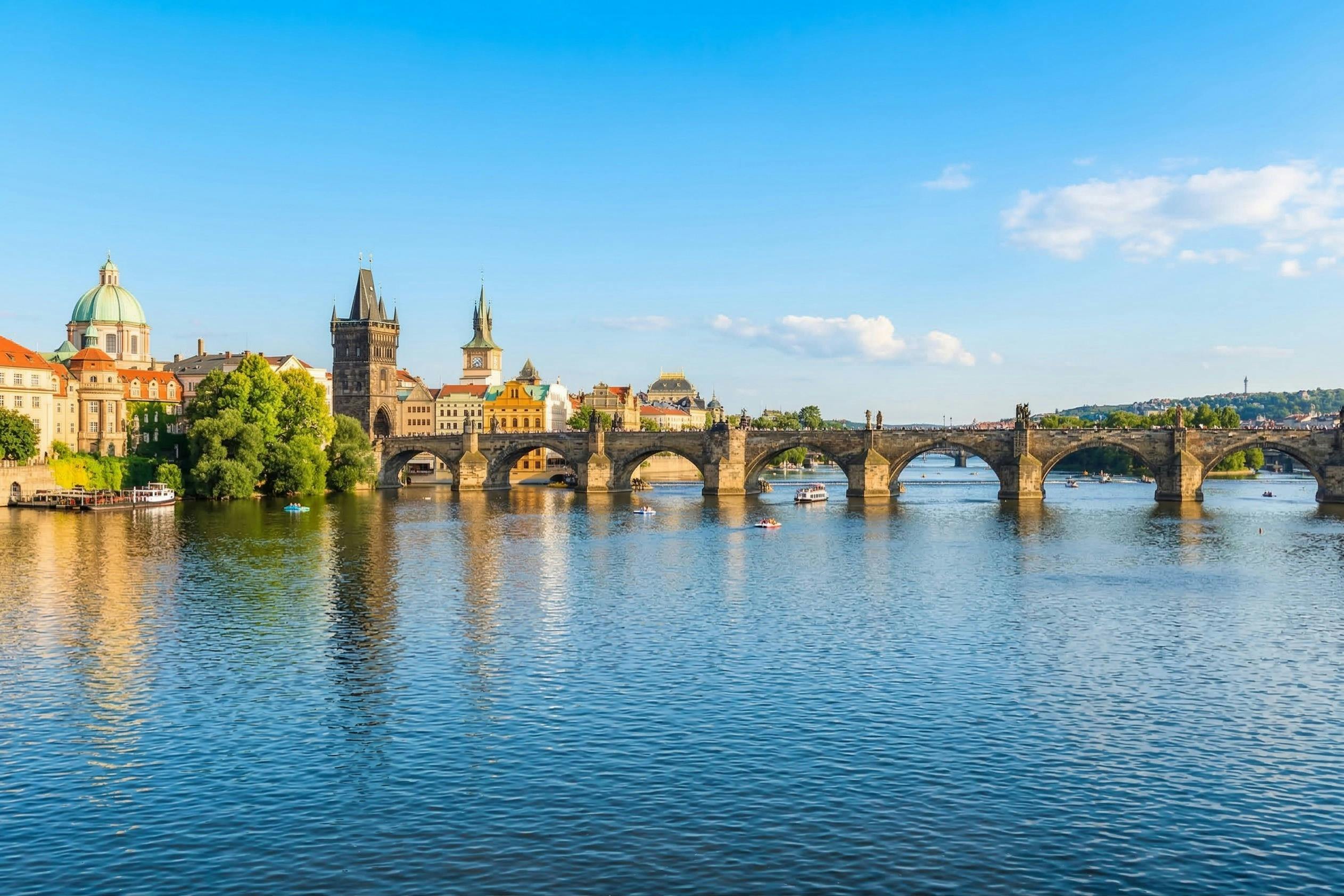 A historic stone bridge spans over a calm river, with buildings and towers in the background under a clear blue sky.