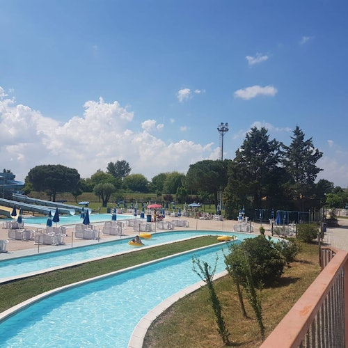 Un parco acquatico con un fiume pigro, sedie a sdraio, ombrelloni, prati verdi e alberi sparsi sotto un cielo parzialmente nuvoloso.