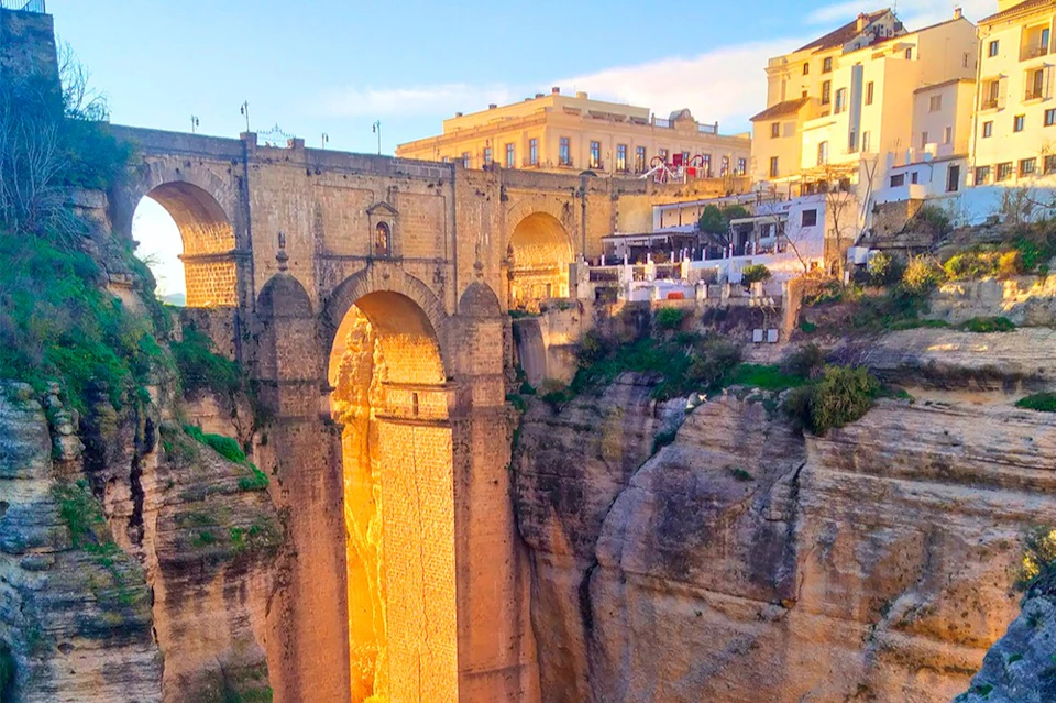 A stone bridge spans a deep gorge with a historic building on the top, illuminated by sunlight under a blue sky.