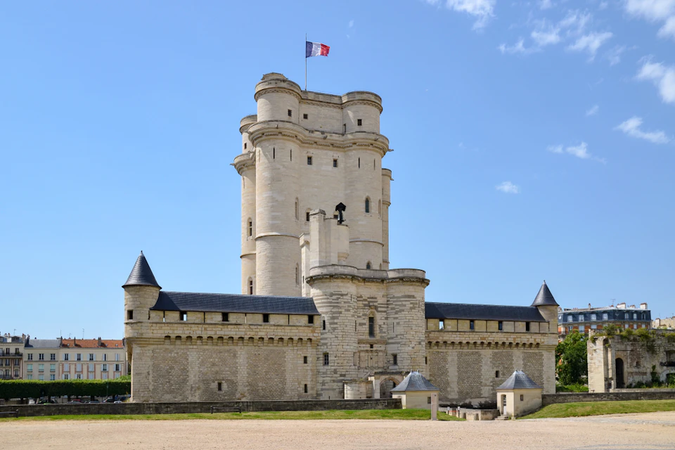 Stone castle with a tall central tower, smaller adjoining structures, and a French flag on top, set against a clear blue sky.
