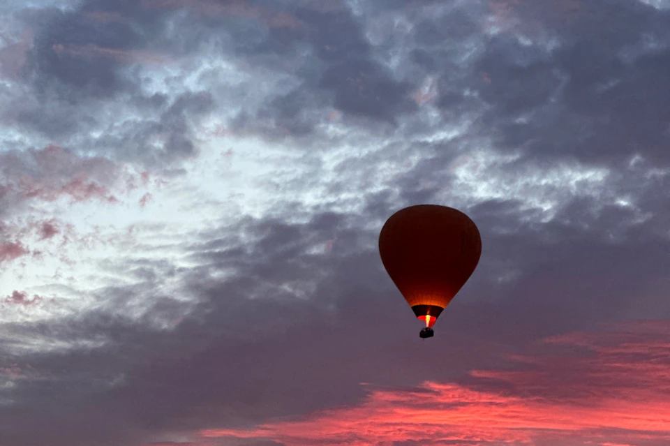 A hot air balloon is silhouetted against a vibrant, cloudy sky at sunset, with a glowing burner beneath the balloon.