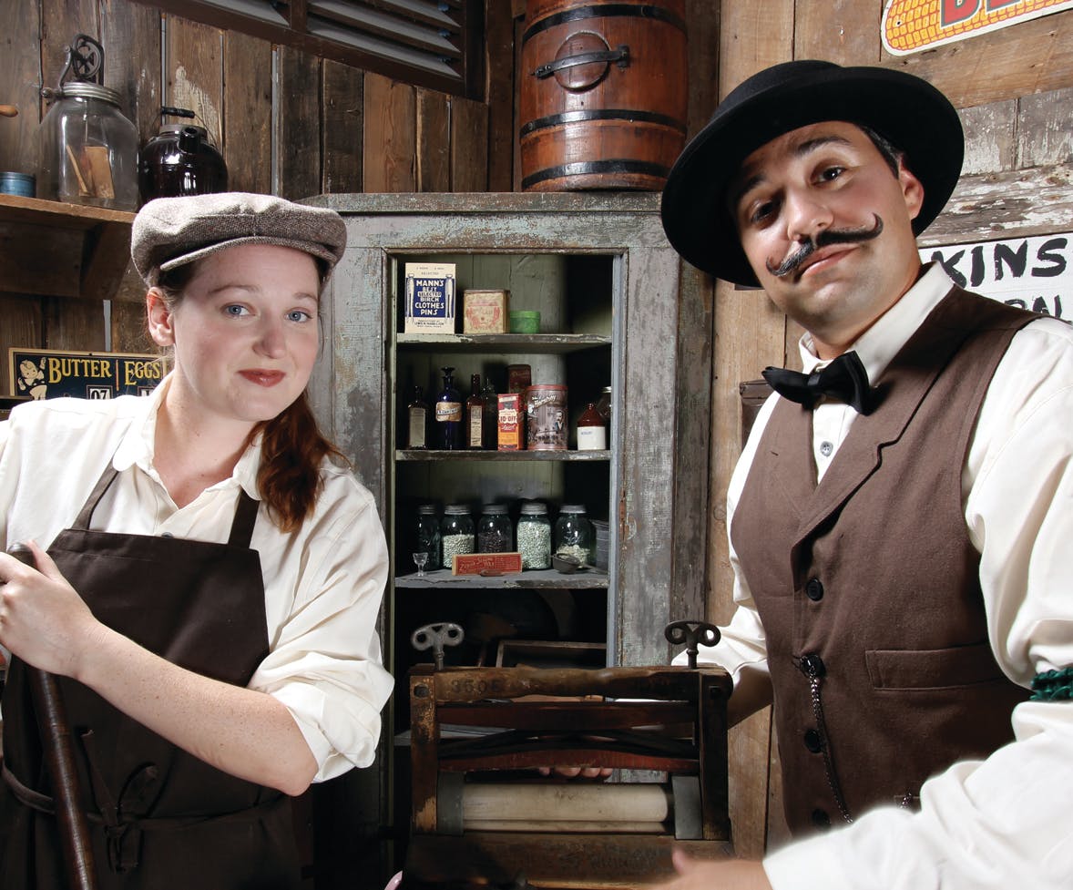 Two people in vintage attire stand in an old-fashioned shop with various goods on shelves behind them.
