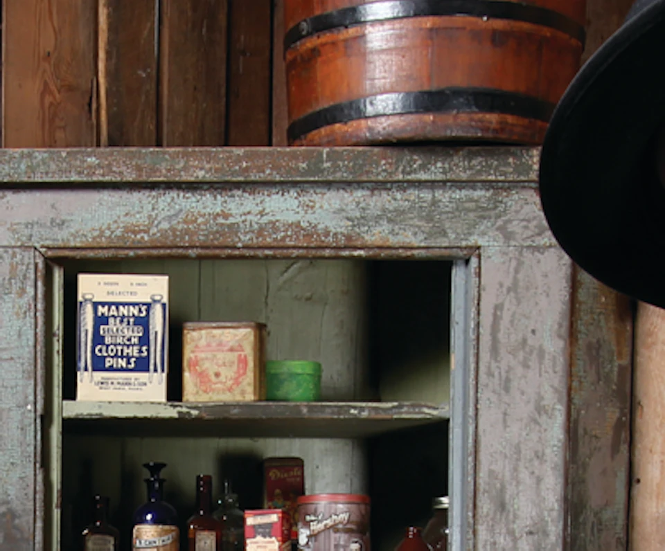 Two people in vintage attire stand in an old-fashioned shop with various goods on shelves behind them.
