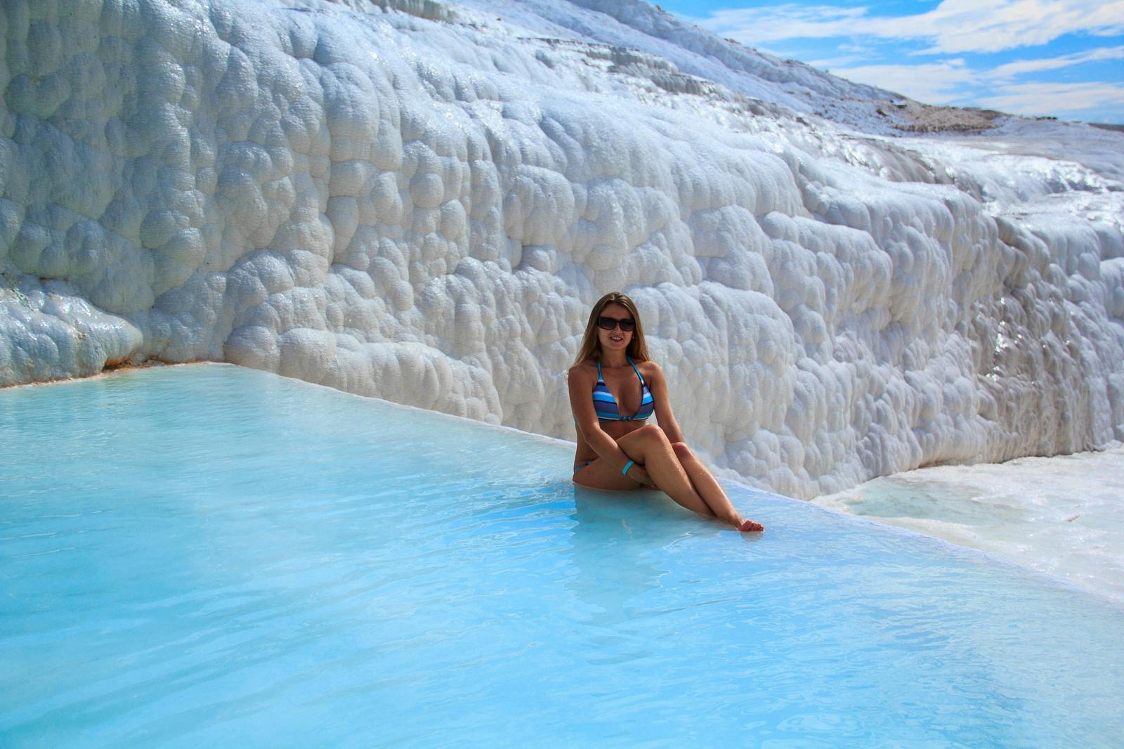 A woman in a bikini sits on the edge of a natural pool with white, terraced rock formations in the background.