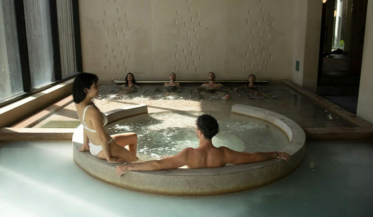Um spa interior sereno, onde as pessoas relaxam numa banheira de hidromassagem e numa piscina maior, rodeado por um interior moderno com iluminação suave.