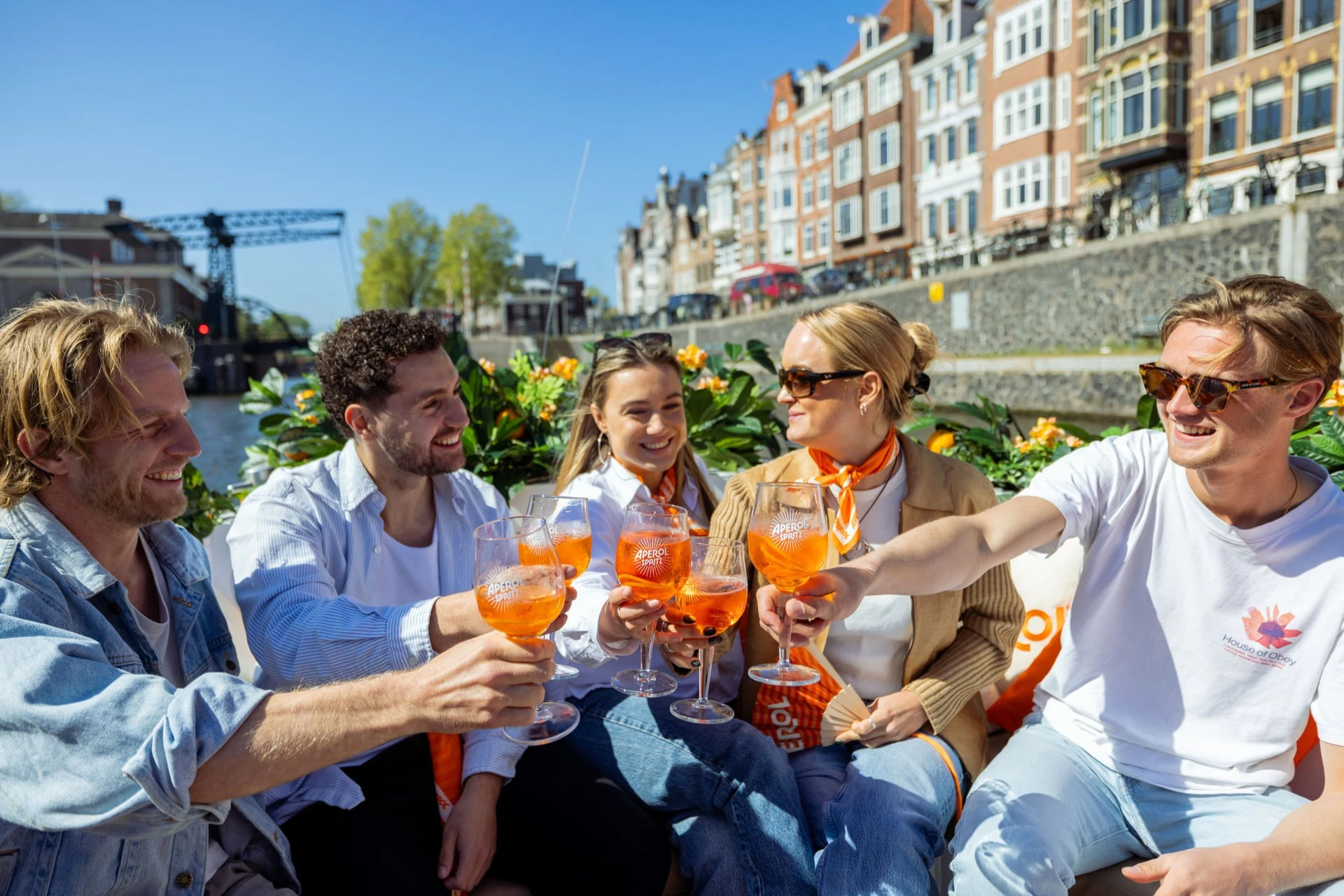 Group of people sitting outdoors, holding glasses with orange drinks, enjoying a sunny day near a canal with buildings.