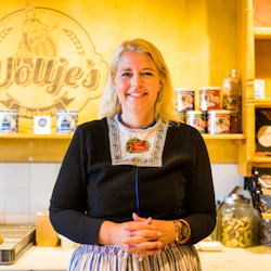 En kvinna i traditionell klädsel står bakom disken i ett bageri med "Woltje's" skrivet på väggen.