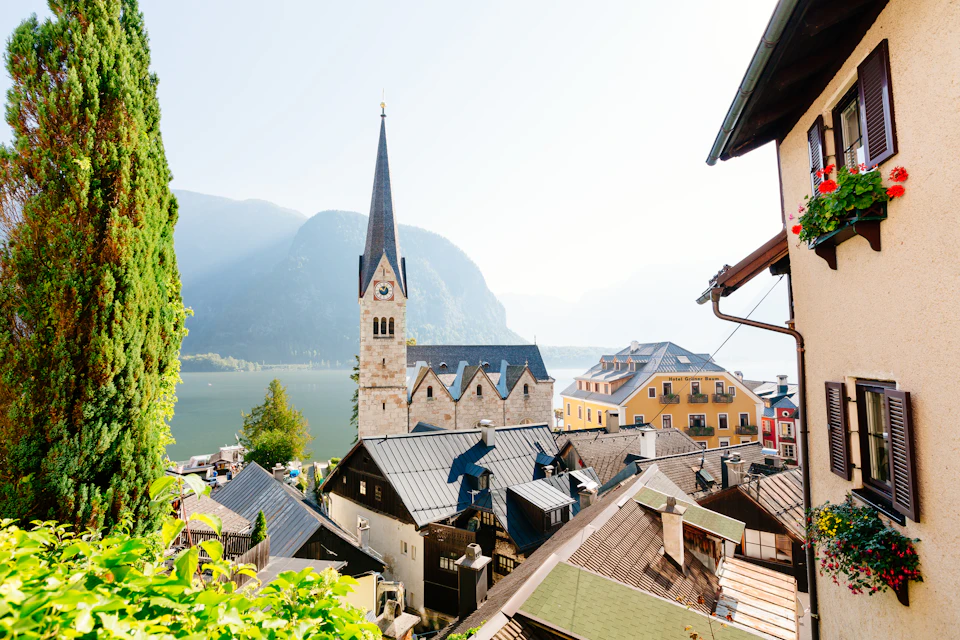 A picturesque village with a prominent church tower, traditional rooftops, and a lake with mountains in the background.