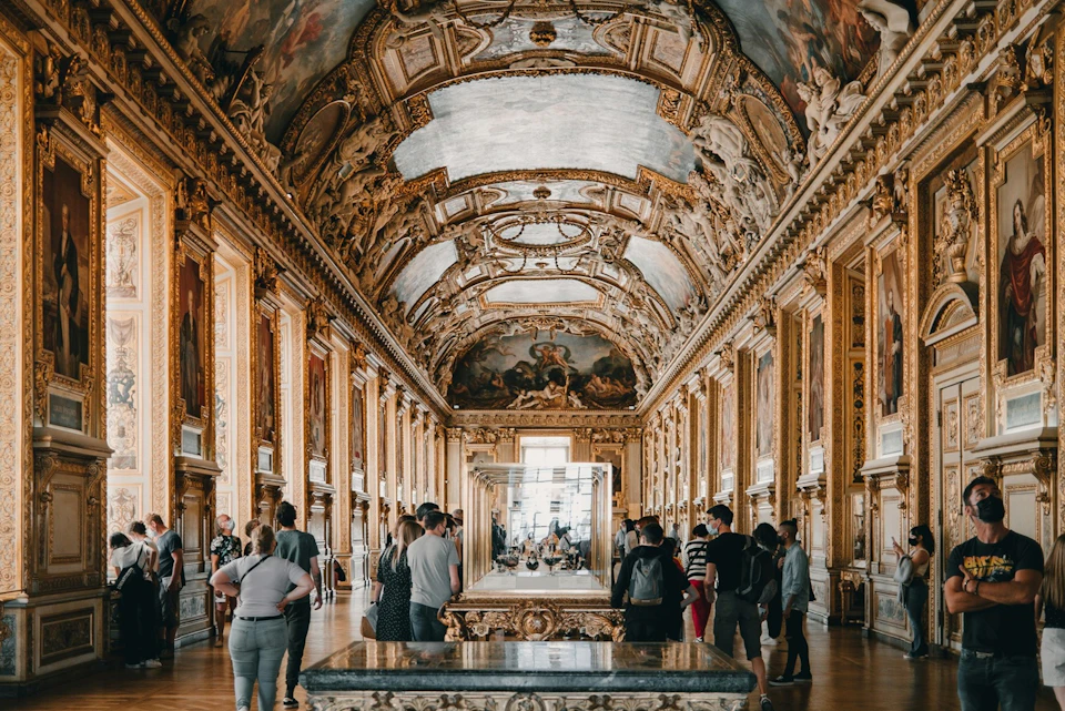 People admire art in a grand gallery with ornate gilded walls, a painted ceiling, and display cases.