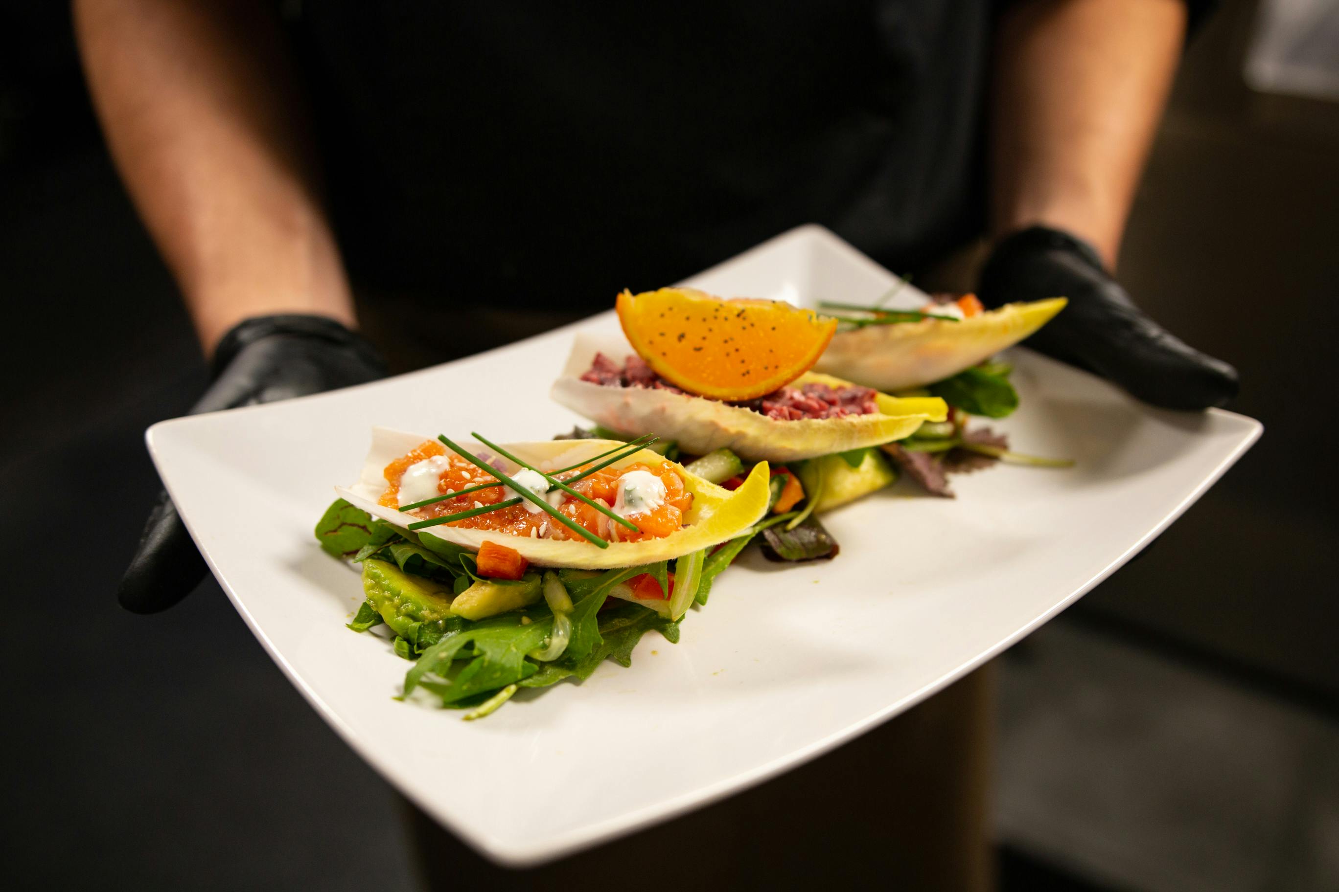A person wearing black gloves holds a plate of gourmet salads garnished with orange slices and fresh greens.