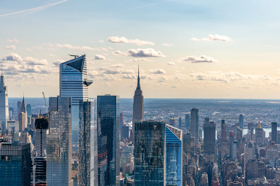 Skyscrapers in New York City with the Empire State Building centered in the background under a partly cloudy sky.