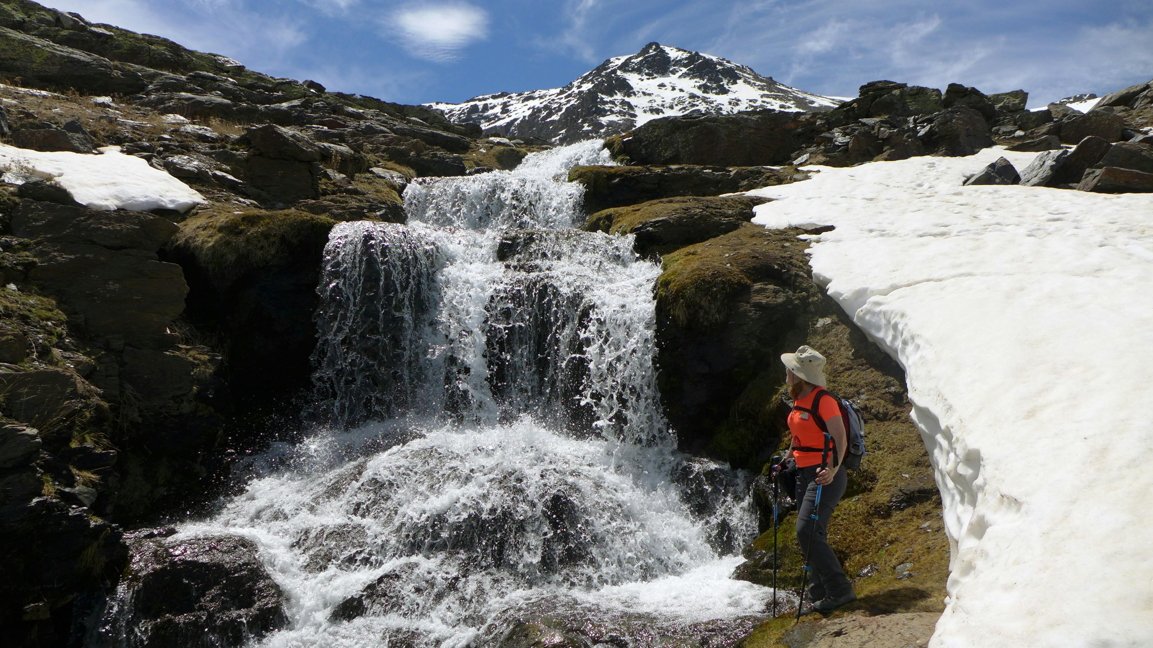 A hiker in a red shirt stands beside a waterfall on a rocky, snow-patched mountain under a blue sky.