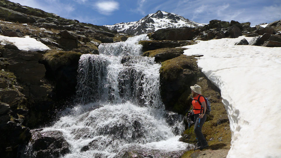 A hiker in a red shirt stands beside a waterfall on a rocky, snow-patched mountain under a blue sky.