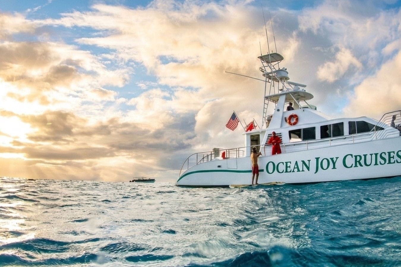 Μια βάρκα που ονομάζεται «Ocean Joy» στον ωκεανό κατά το ηλιοβασίλεμα, με ανθρώπους στο σκάφος και ένα άτομο να στέκεται σε μια σανίδα δίπλα της.
