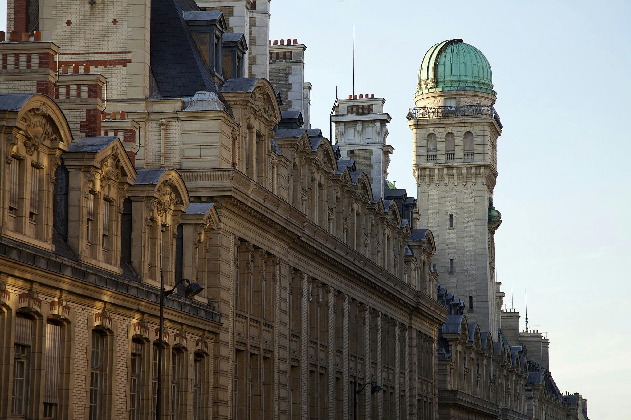 Ornate, historic buildings with detailed facades and mansard roofs line a street, with a tall tower adorned with a green dome.