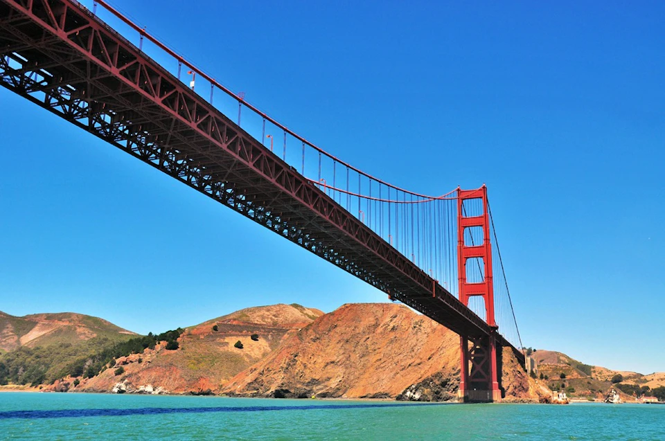 Red suspension bridge over turquoise water with rocky hills in the background under a clear blue sky.
