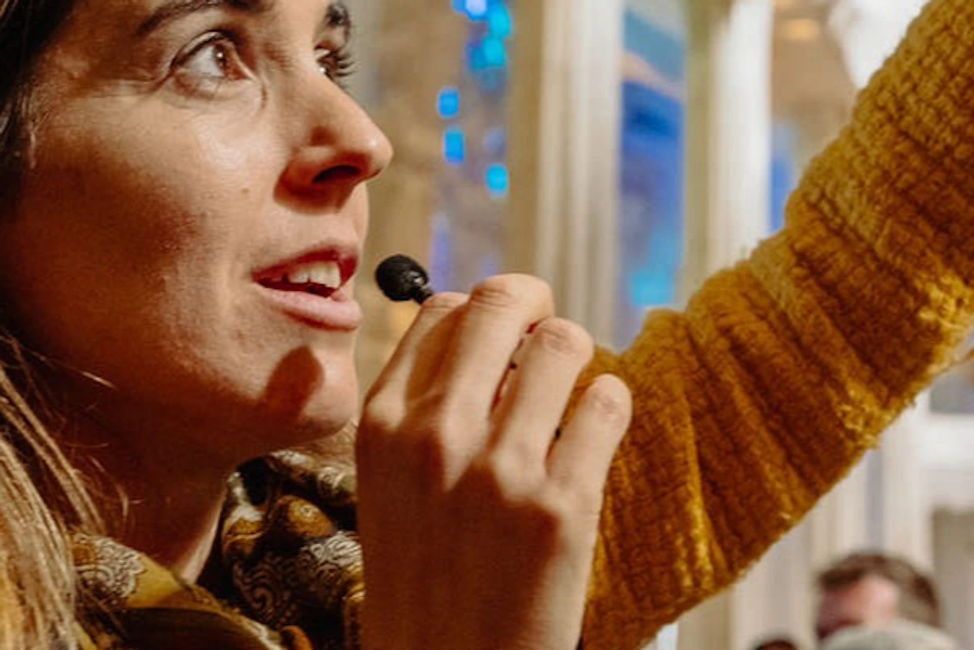 A woman in a yellow jacket speaks into a microphone while pointing upwards in a cathedral with stained glass windows.