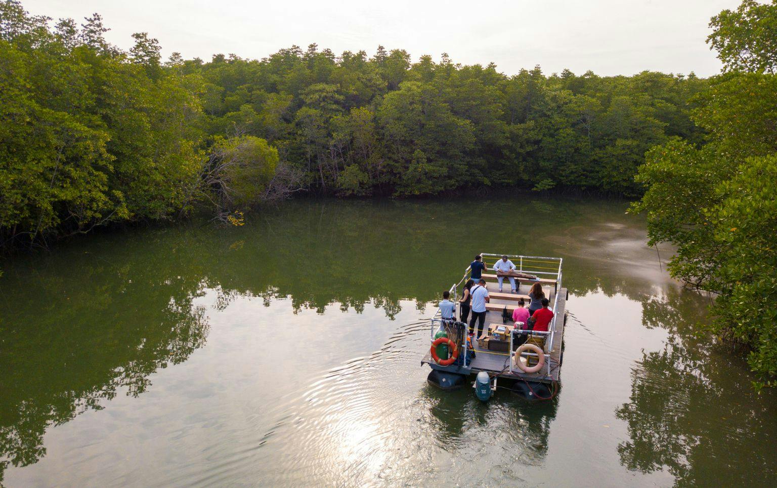 A small boat with people on board is floating on calm water, surrounded by dense greenery and trees under a cloudy sky.