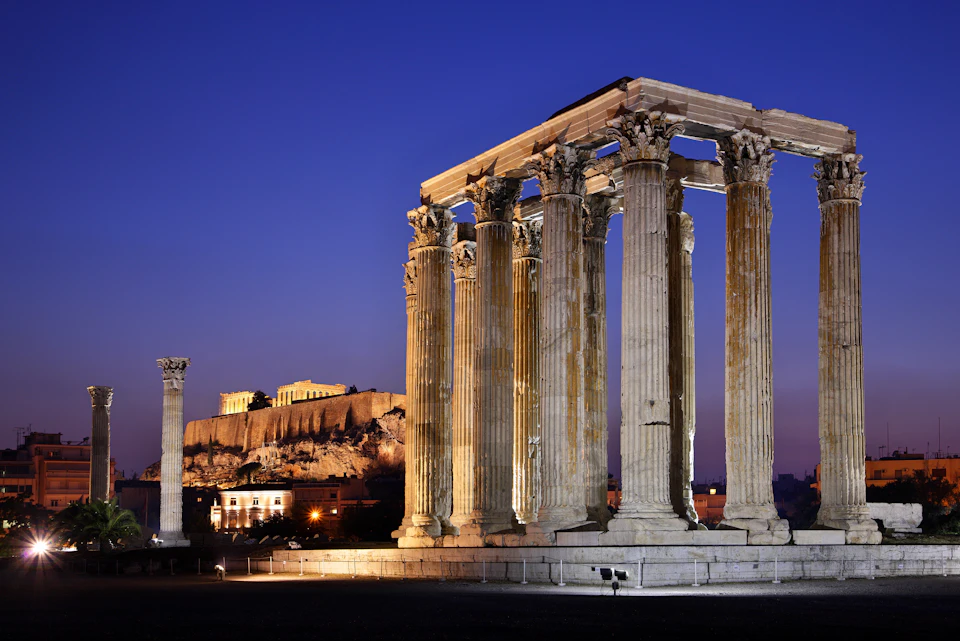 Ancient Greek temple ruins with tall columns, the illuminated Acropolis in the background under a deep blue evening sky.