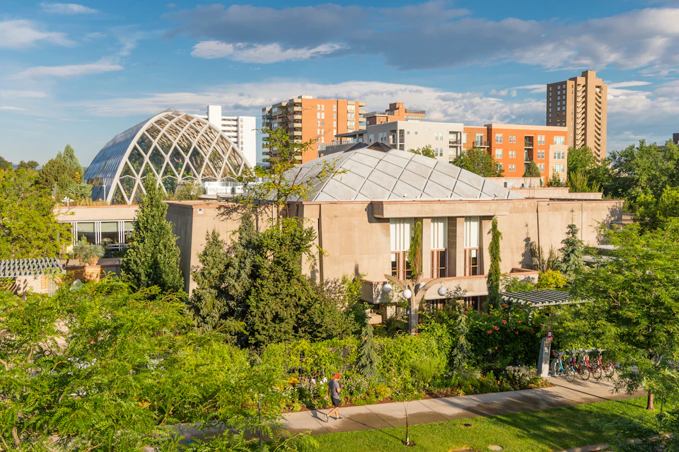 A building with a geometric dome roof, lush greenery in the foreground, and several modern buildings in the background.