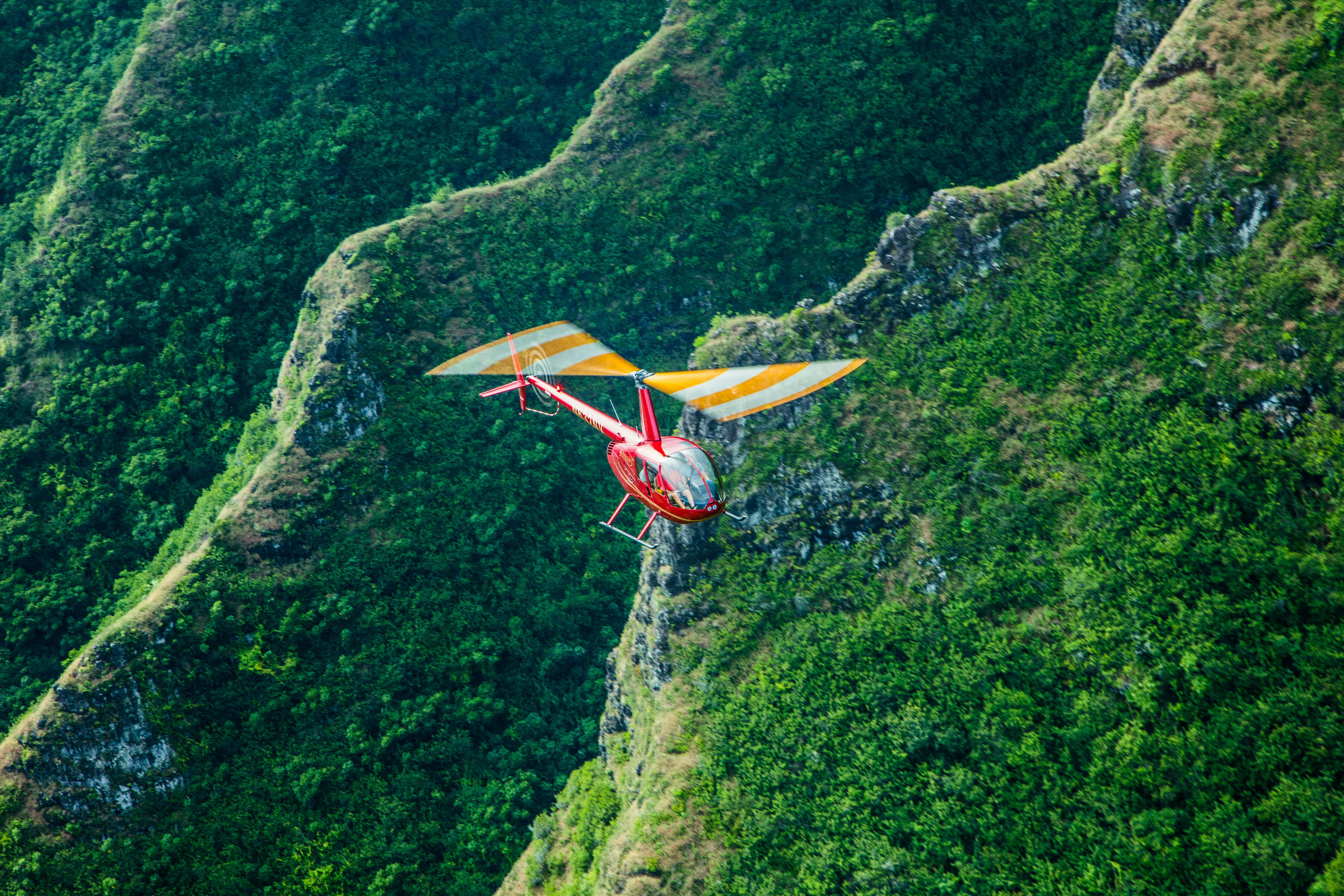 A red helicopter with yellow-striped blades flies over a lush, green, hilly landscape.