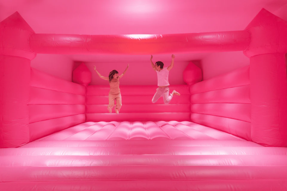 Two people jump inside a large, pink, inflatable bouncy house with padded walls and floor.