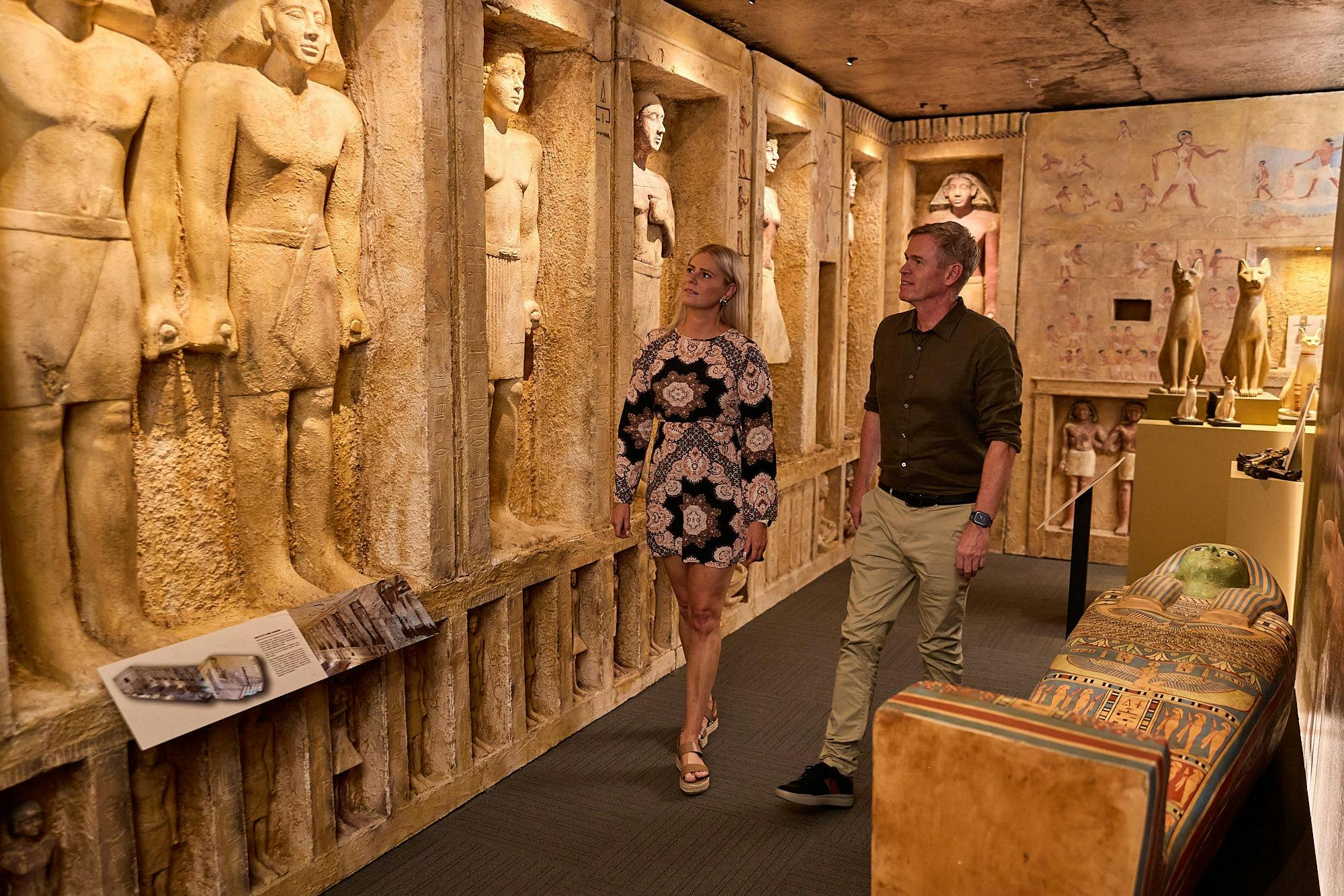 Dva lidé procházejí muzejní expozicí se sochami a řezbami na zdech ve starověkém egyptském stylu.