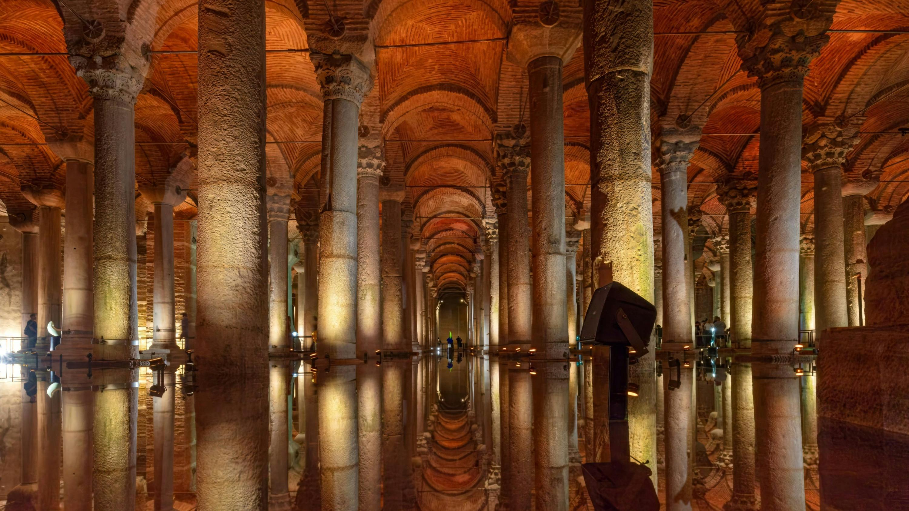 An illuminated underground stone structure with tall columns and arches reflected on a shallow pool of water.