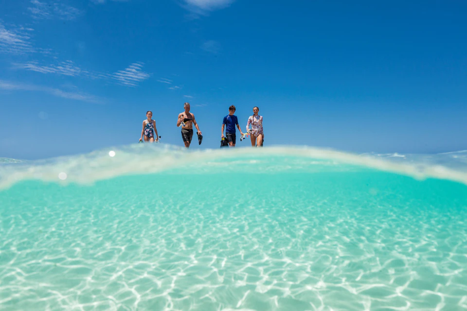 Four people in swimwear wading through clear turquoise water under a bright blue sky.