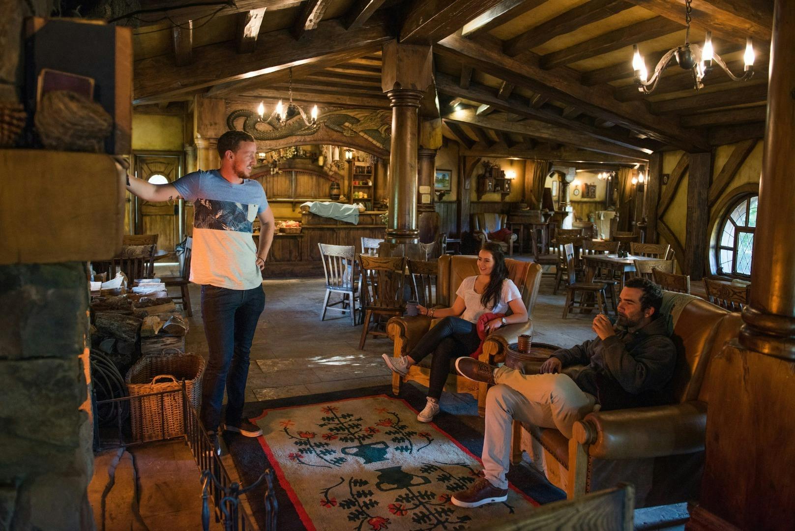Three people converse in a cozy, rustic tavern with wooden beams, chairs, tables, and warm ambient lighting.