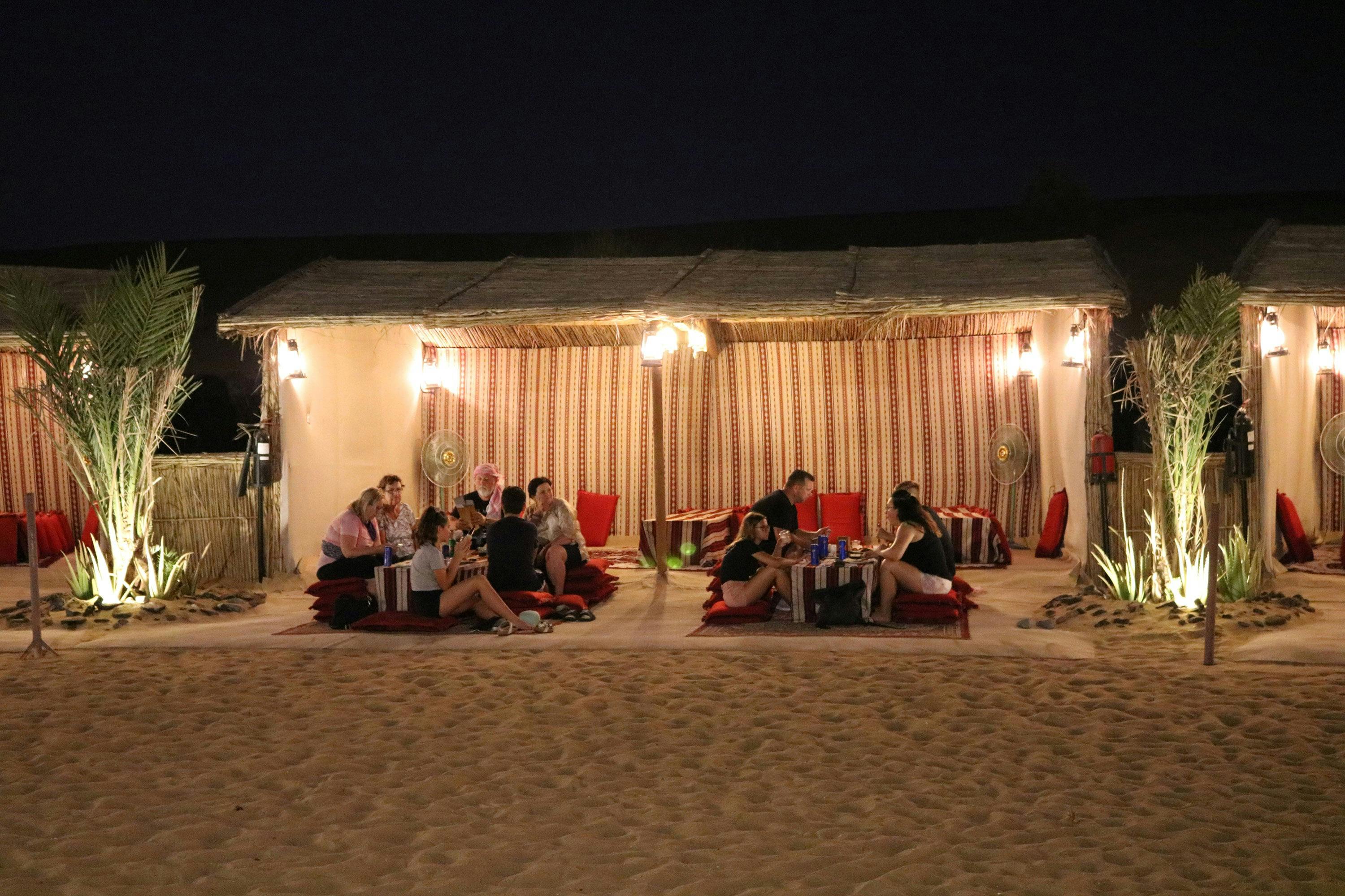 Groups of people seated on cushions under a canopy, dining and socializing at night in a desert setting.