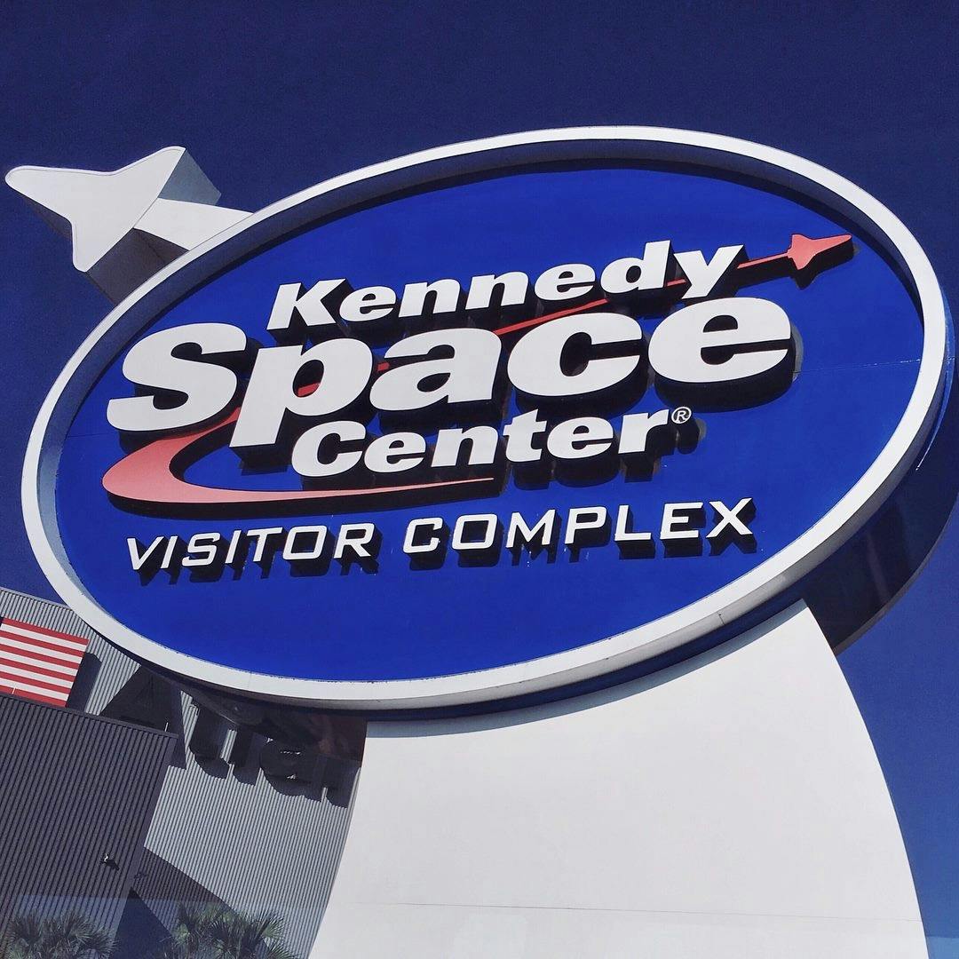 Kennedy Space Center Visitor Complex sign with a rocket icon, set against a blue sky background.