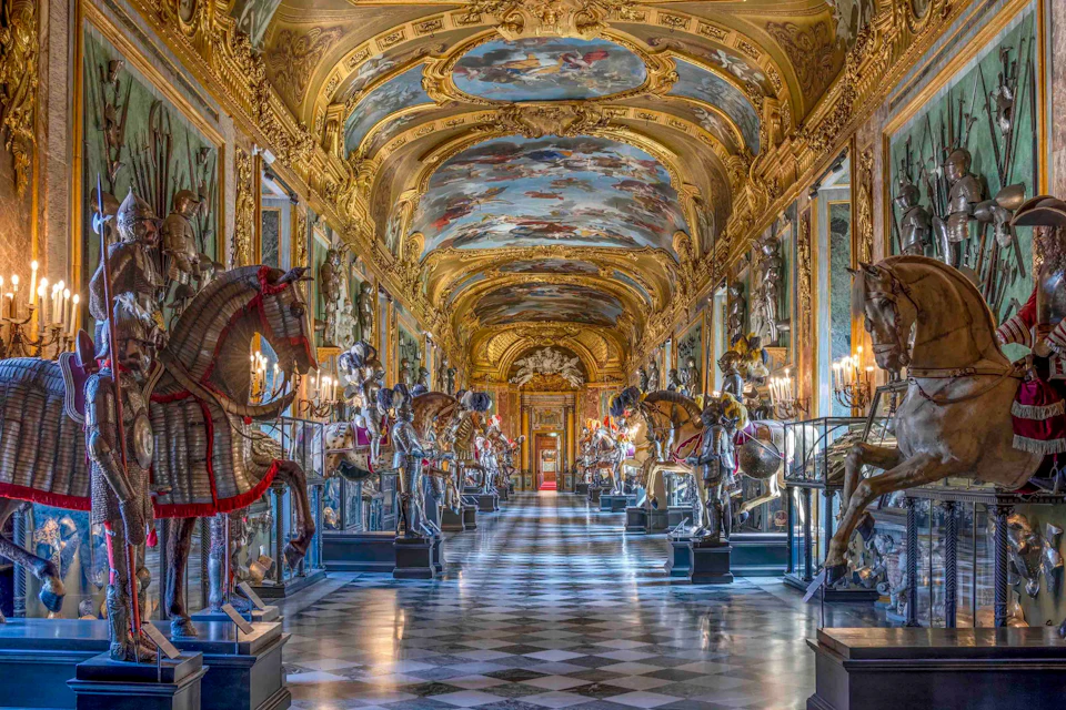 Ornate gallery with a painted vaulted ceiling, suits of armor, and decorated horses along a checkered floor.