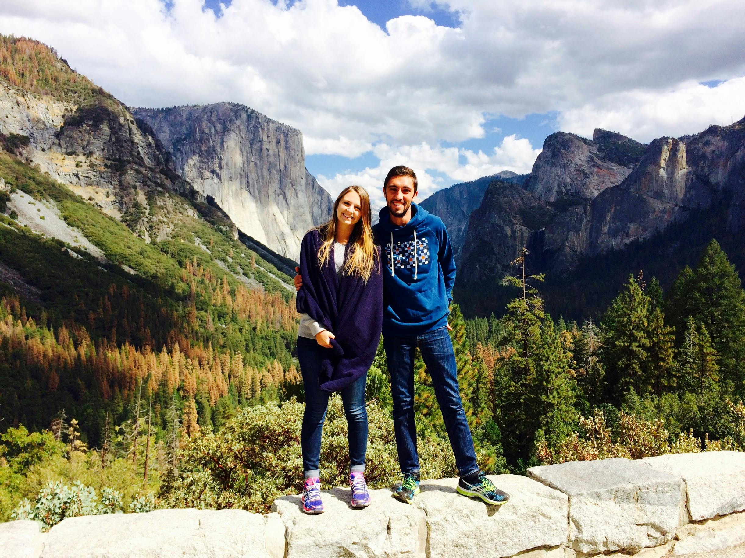 一对微笑的夫妇站在石架上，背景是风景秀丽的山景和多云的天空。