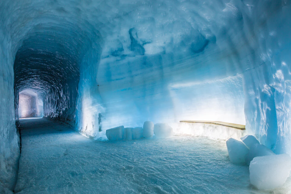 Ice cave with blue-toned walls, chunks of ice on the floor, and a wooden handrail along the side, illuminated by artificial light.
