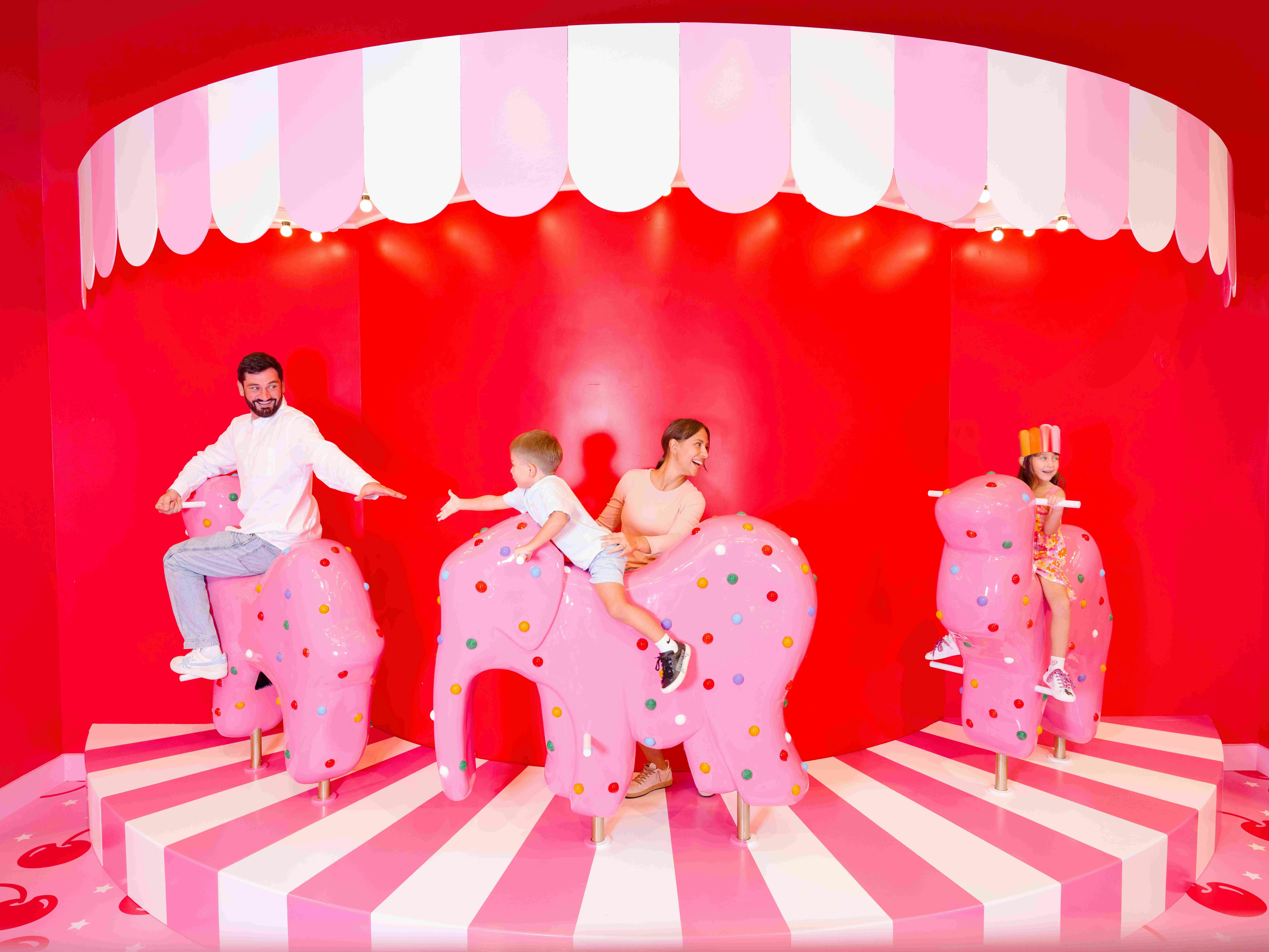 Tři lidé jezdí na růžových, puntíkovaných sloních sedadlech v pestrobarevném cirkusovém prostředí s červeným a růžovým dekorem.