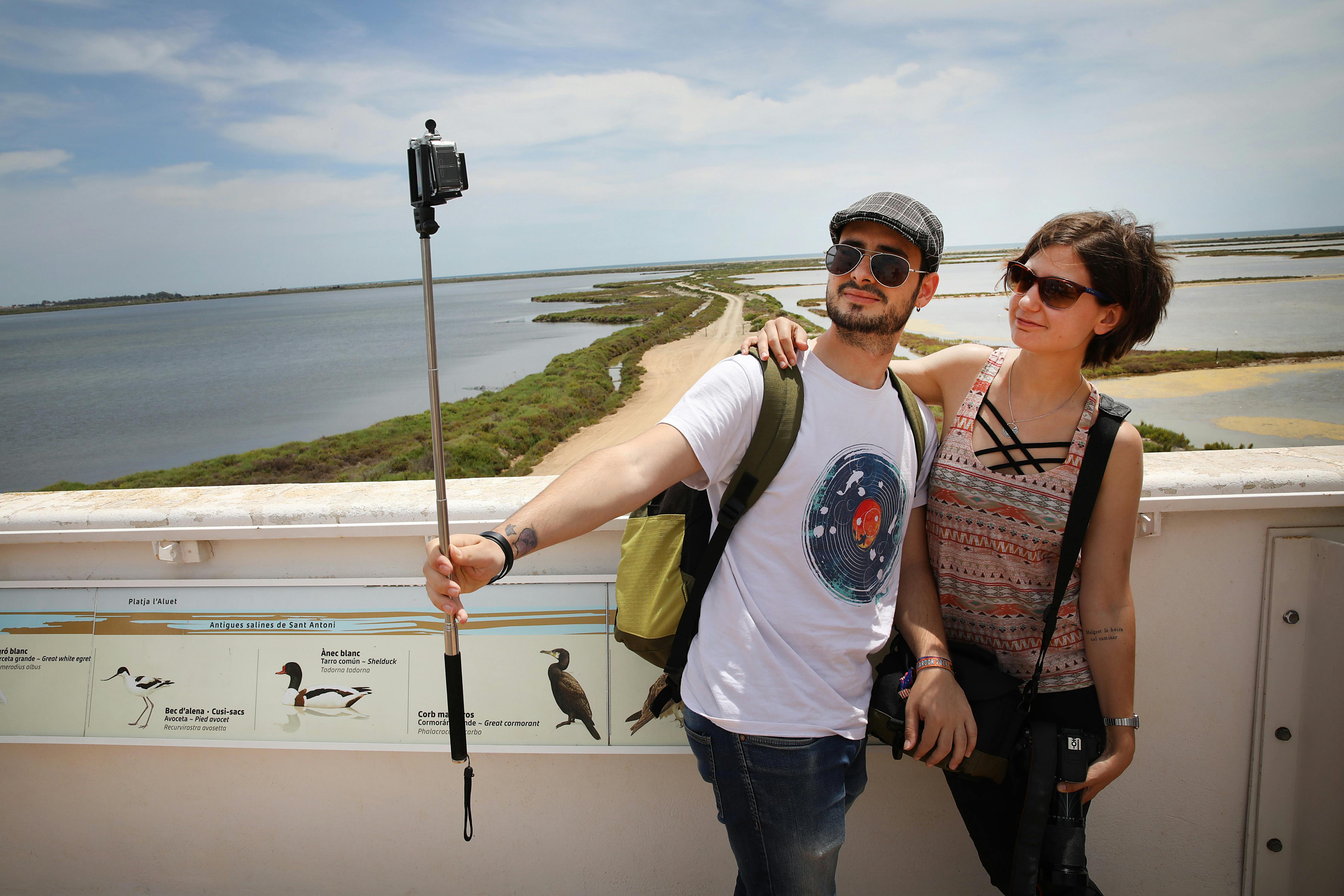 Dva lidé, kteří si dělají selfie na mostě s výhledem na malebnou bažinu, na sobě mají ležérní oblečení a sluneční brýle.