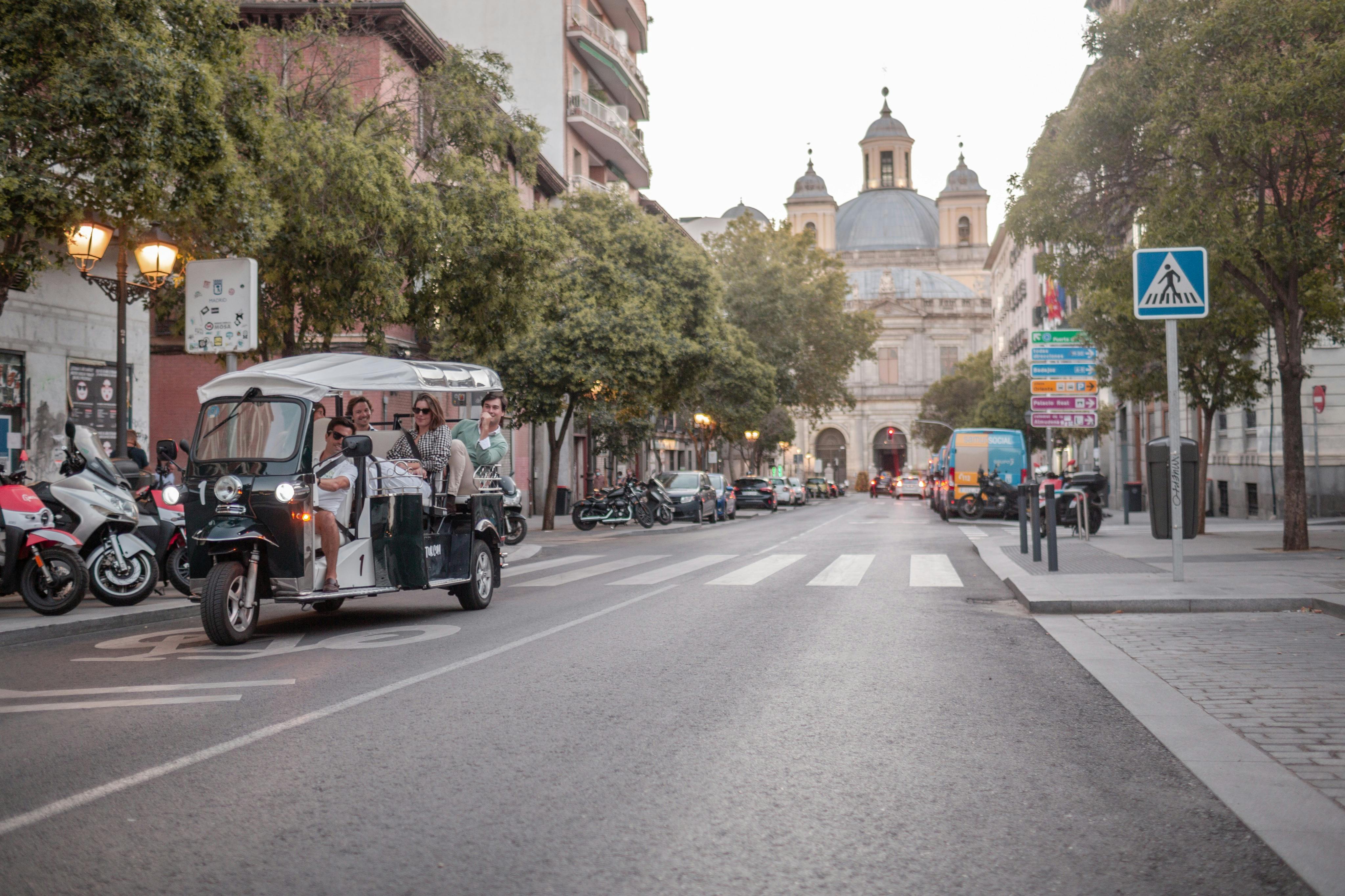 A tuk-tuk drives down a street lined with trees, buildings, and parked vehicles, with a domed building in the background.