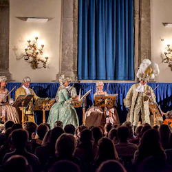 Musiker i genomarbetade periodkostymer som uppträder på scenen med blå gardiner, tända lampetter och en full publik som tittar på.