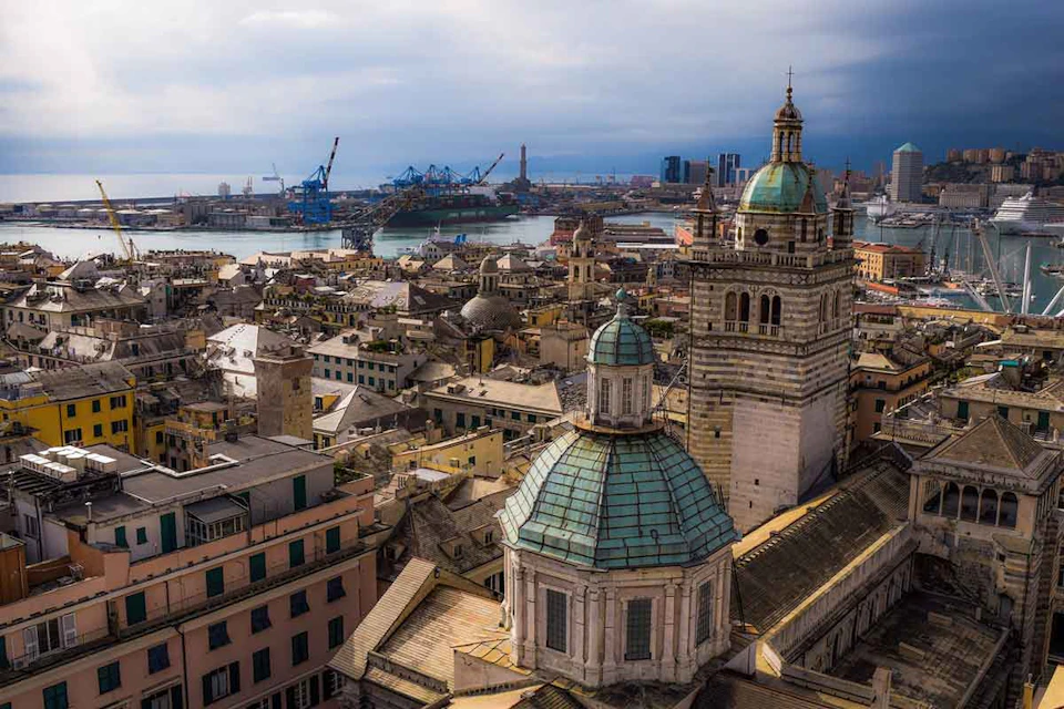 Aerial view of a city with historic buildings, green-domed church towers, and a distant port with cranes and ships.