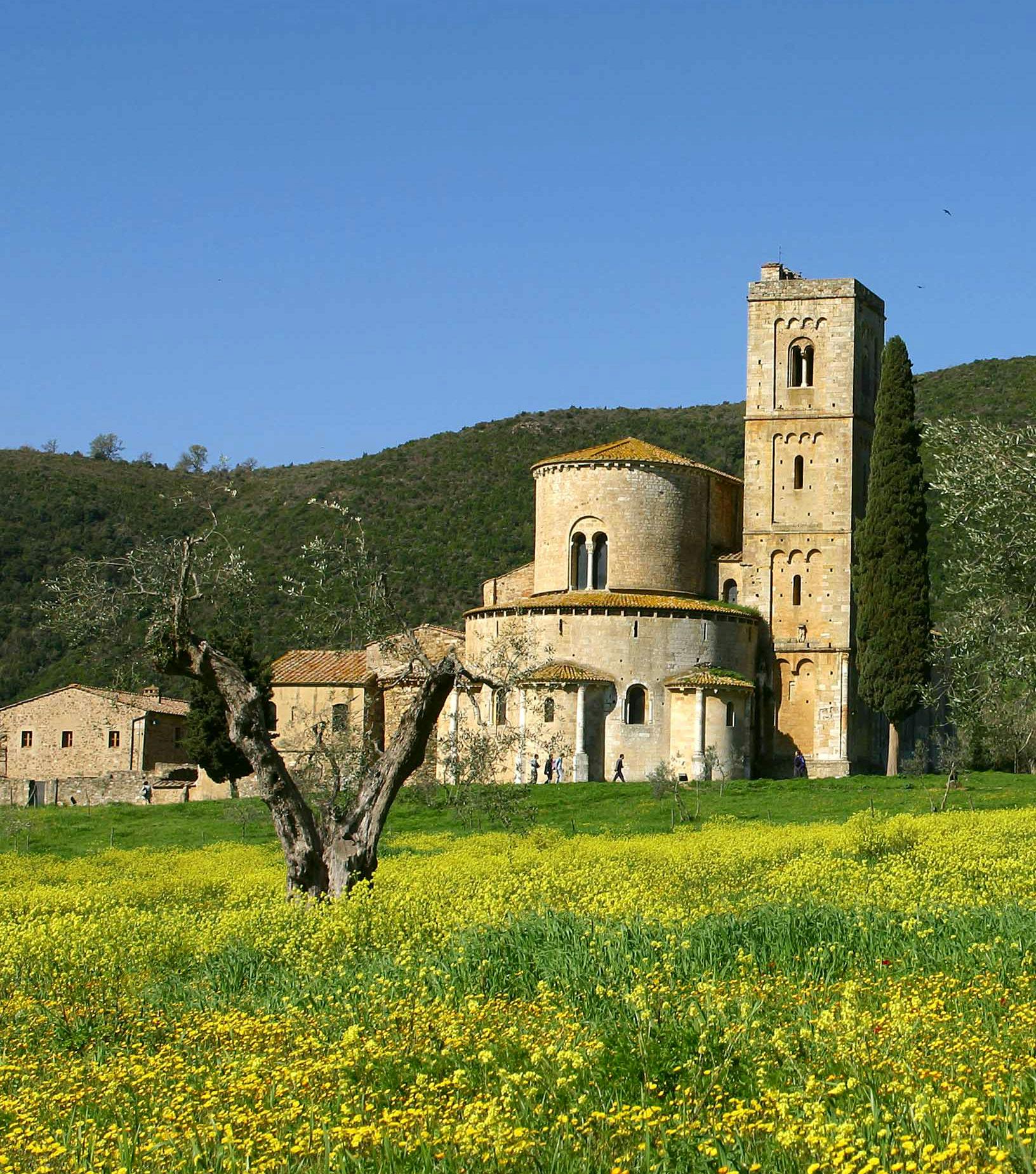 A historic stone church with a tall tower and rounded apse, nestled in a meadow with yellow flowers, against a backdrop of green hills.
