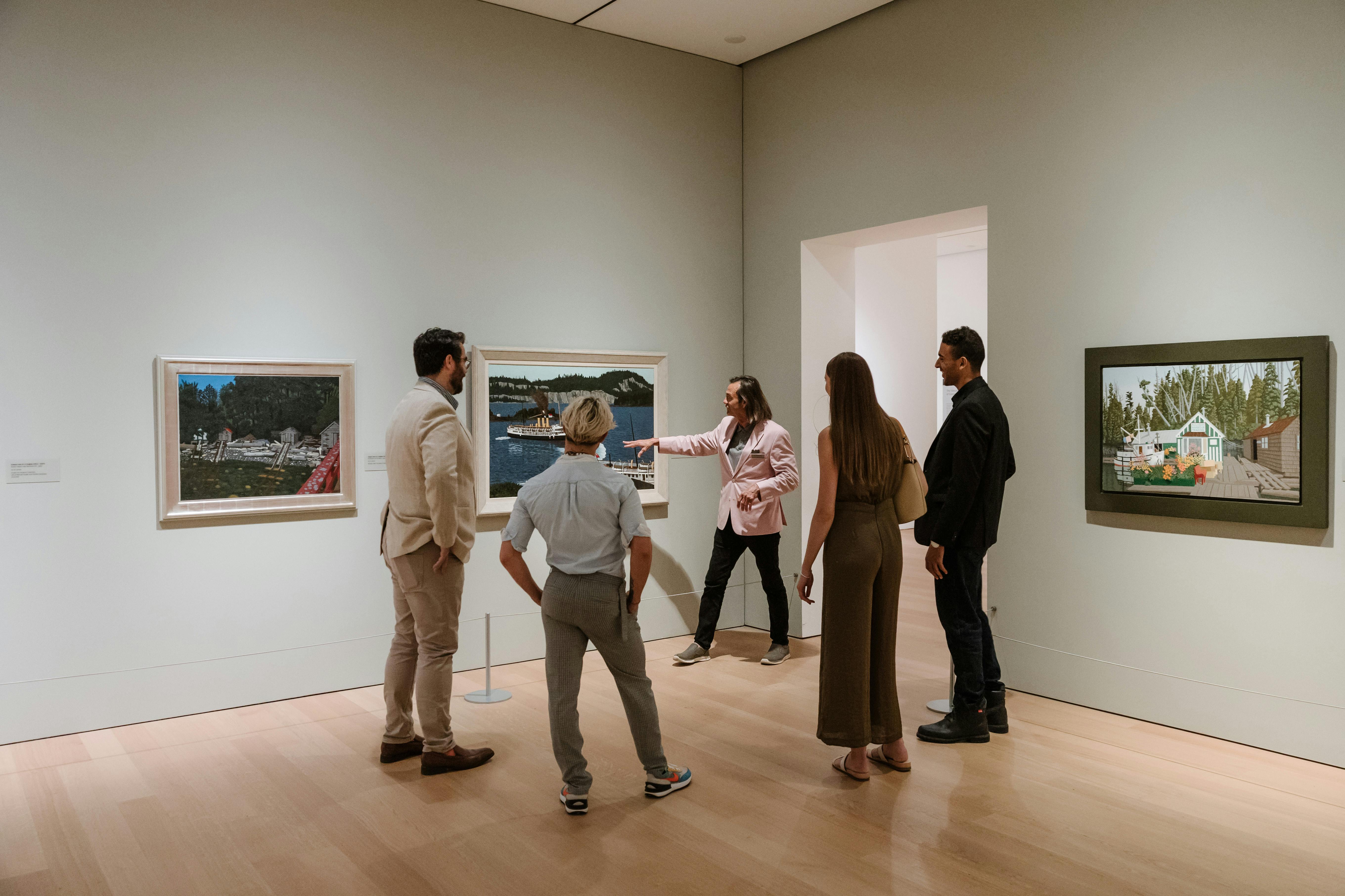 Un groupe de personnes admire des tableaux dans une galerie d'art. Une personne vêtue d'une veste rose fait un geste en direction d'une peinture représentant une scène côtière.